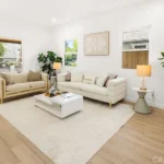 Living room with light sofa, chair, coffee table, plants, big windows, and wood floor