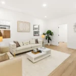 Living room with beige sofa, white coffee table, plants, lamps, and white walls