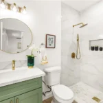 Bathroom with round mirror, green sink vanity, toilet, shower, and plants