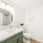 Bathroom with round mirror, white counter, green cabinet, toilet, and small window