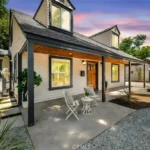 A charming two-story house with a gabled roof, front porch, and large windows. Two white chairs and a small table are on the porch. The landscaping features gravel, plants, and a pathway leading to the entrance. Trees are visible in the background under a colorful sky.