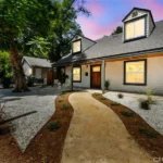 California real estate house with wood walls, dark roof, round path, gravel yard, and plants