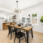 Dining room with wood table, four black chairs, hanging light, plant, and kitchen in back
