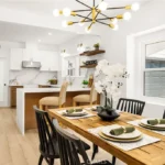 Dining area with wood table set for meal, black chairs, orchid vase, and white kitchen in back