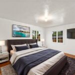 Bedroom with big bed, dark bedding, two lamps, beach wall art, and big windows