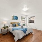 Bedroom with blue white bed, pillows, two lamps, wall art, wood floor, and windows