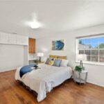 Bedroom with big bed, soft linens, lamp, plants, window, and wood floor