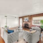 A spacious living room featuring two gray tufted armchairs, a coffee table in the center, and a stone fireplace. There are large windows allowing natural light in and a green potted plant near the window. The walls have a mix of white and wooden paneling.