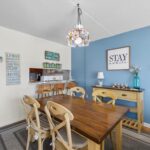 Dining room with wood table, six chairs, blue wall, stay awhile sign, and lamp